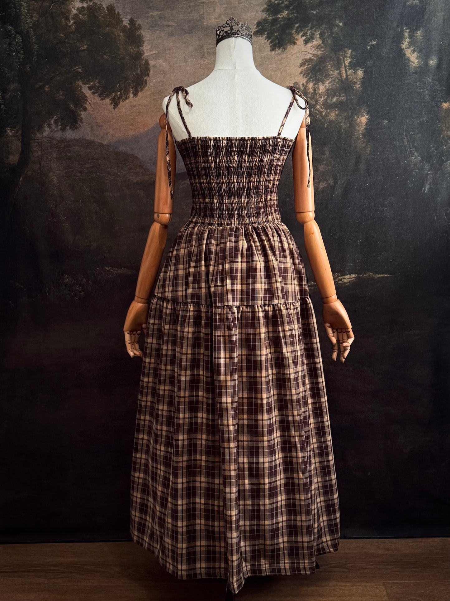 A mannequin wearing a historical fashion inspired brown and beige tartan plaid maxi dress with tie straps against a dark art history background