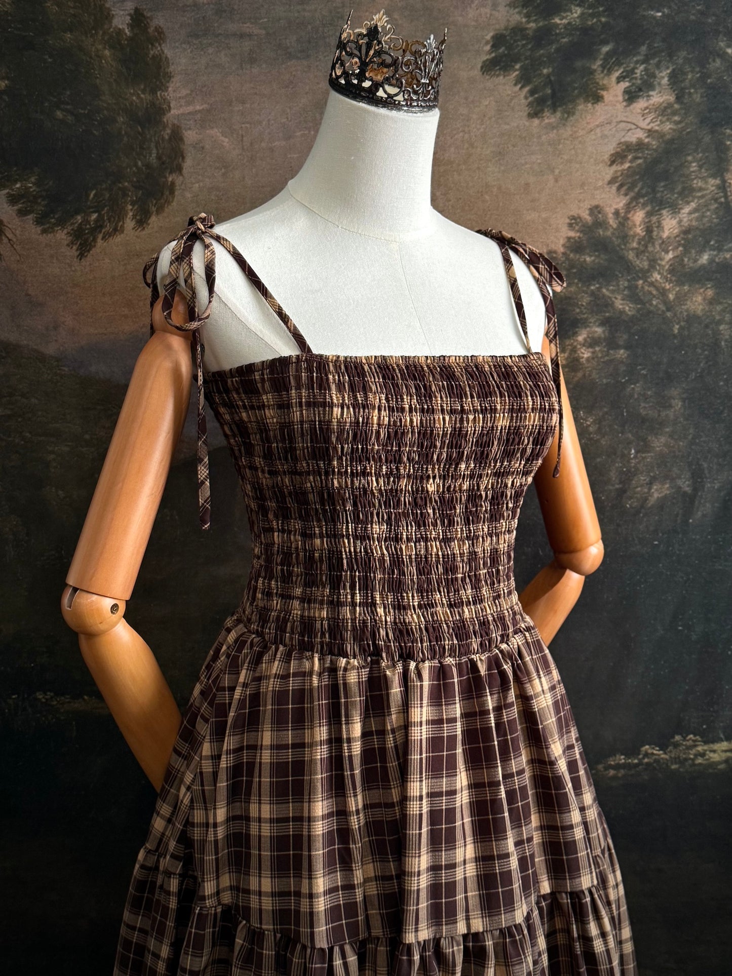A mannequin wearing a historical fashion inspired brown and beige tartan plaid maxi dress with tie straps against a dark art history background