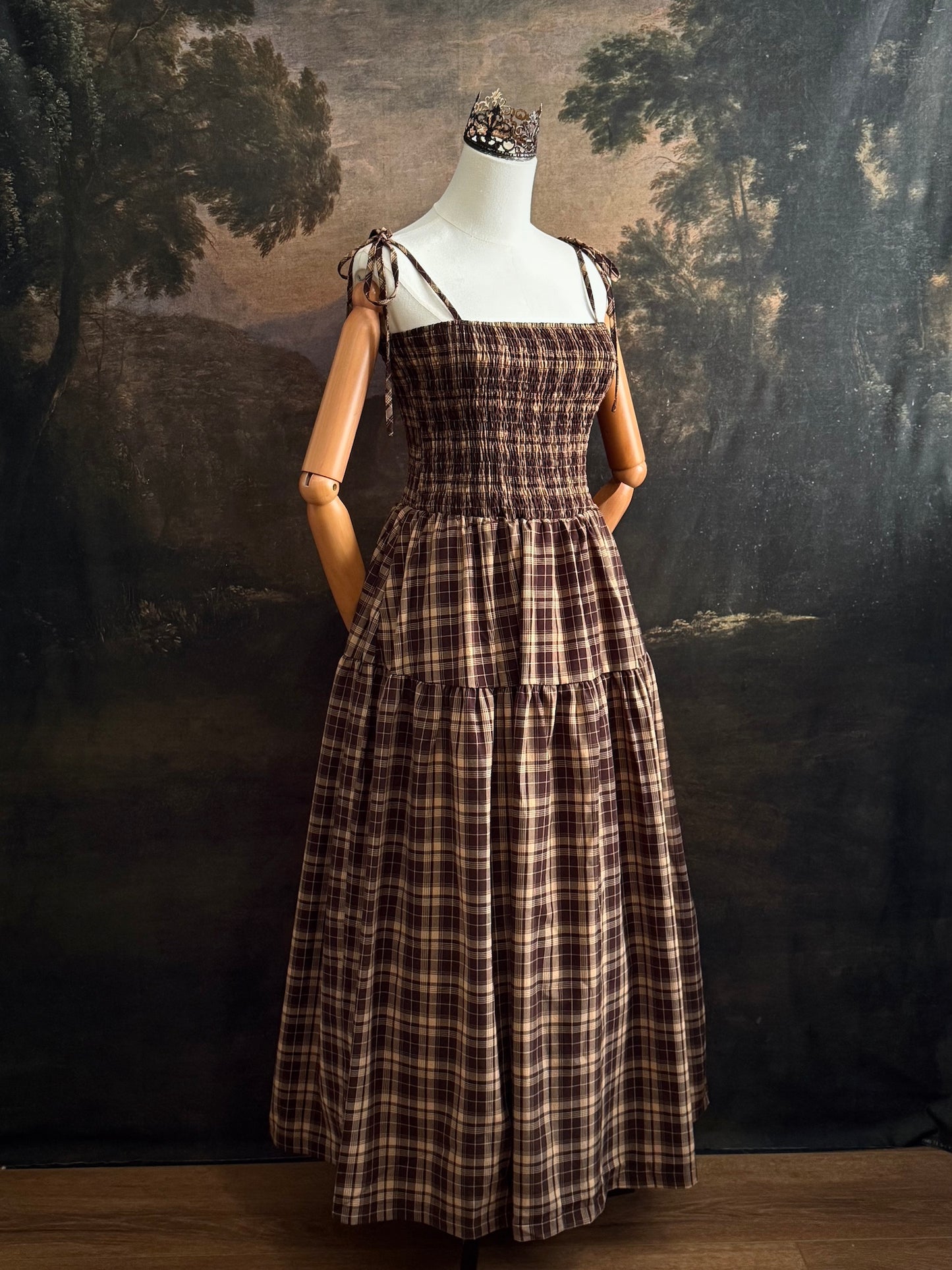 A mannequin wearing a historical fashion inspired brown and beige tartan plaid maxi dress with tie straps against a dark art history background