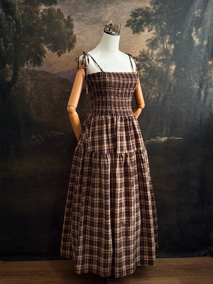A mannequin wearing a historical fashion inspired brown and beige tartan plaid maxi dress with tie straps against a dark art history background