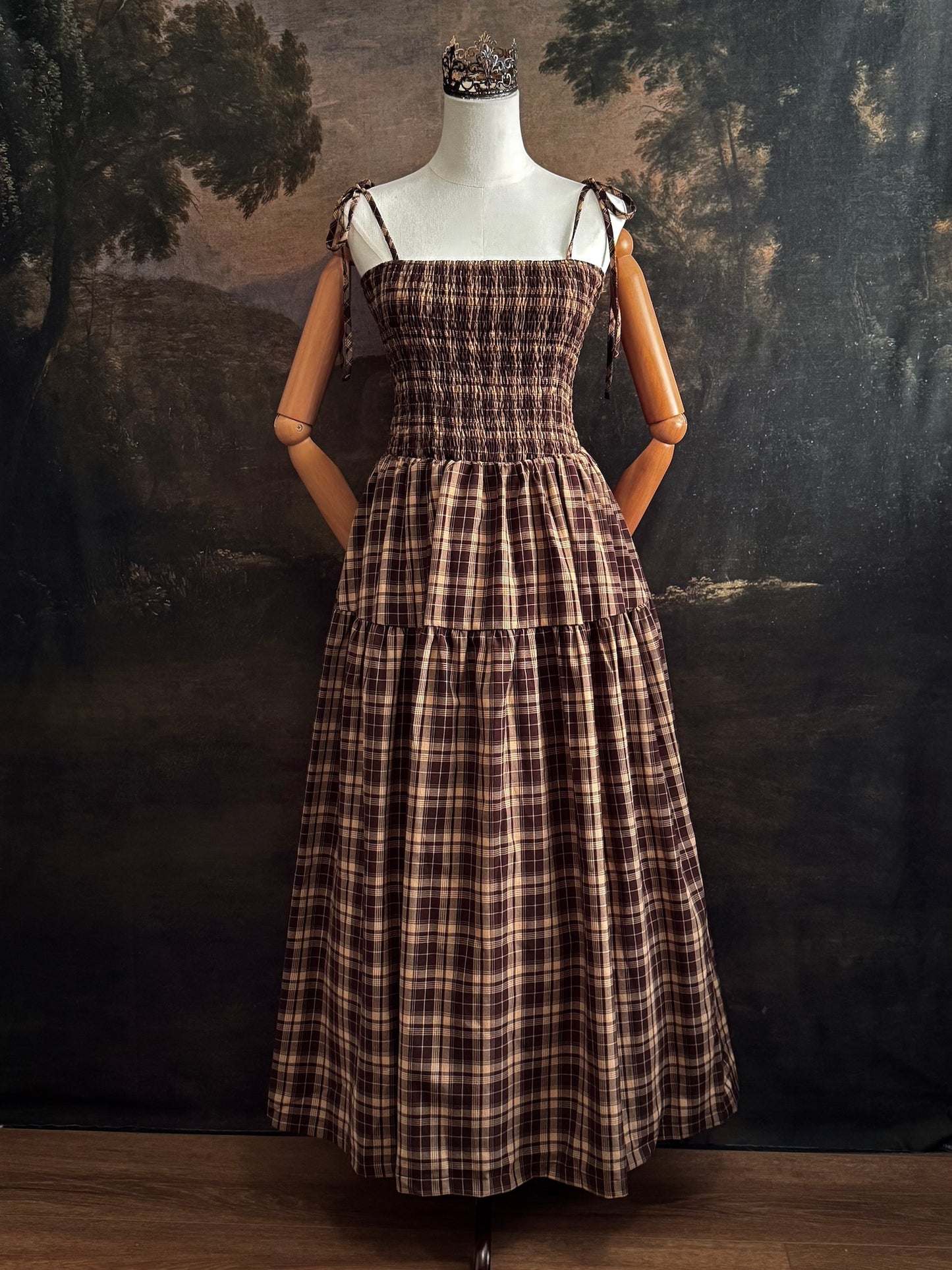 A mannequin wearing a historical fashion inspired brown and beige tartan plaid maxi dress with tie straps against a dark art history background