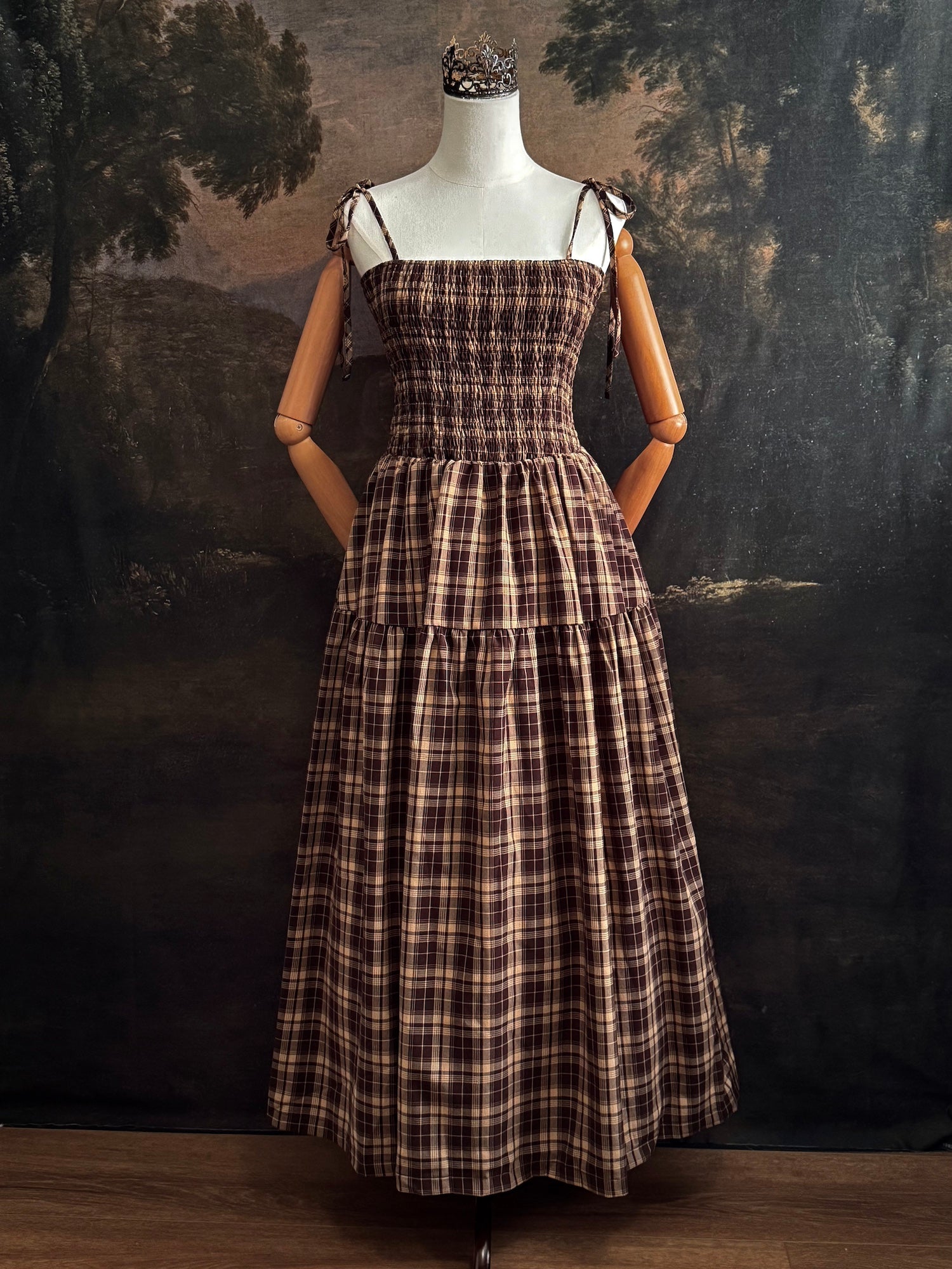 A mannequin wearing a historical fashion inspired brown and beige tartan plaid maxi dress with tie straps against a dark art history background