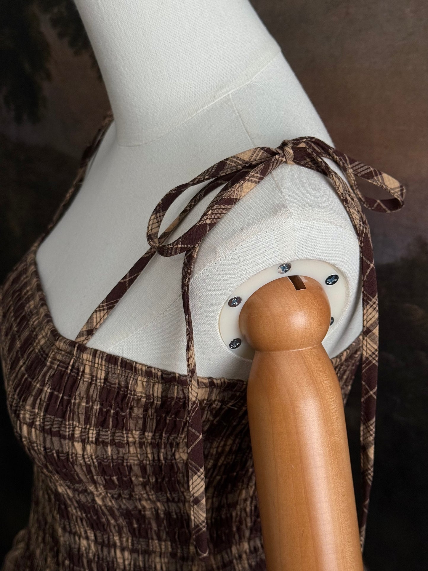 A mannequin wearing a historical fashion inspired brown and beige tartan plaid maxi dress with tie straps against a dark art history background