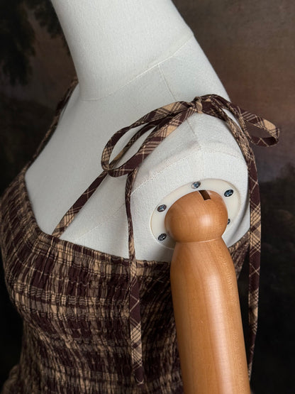 A mannequin wearing a historical fashion inspired brown and beige tartan plaid maxi dress with tie straps against a dark art history background