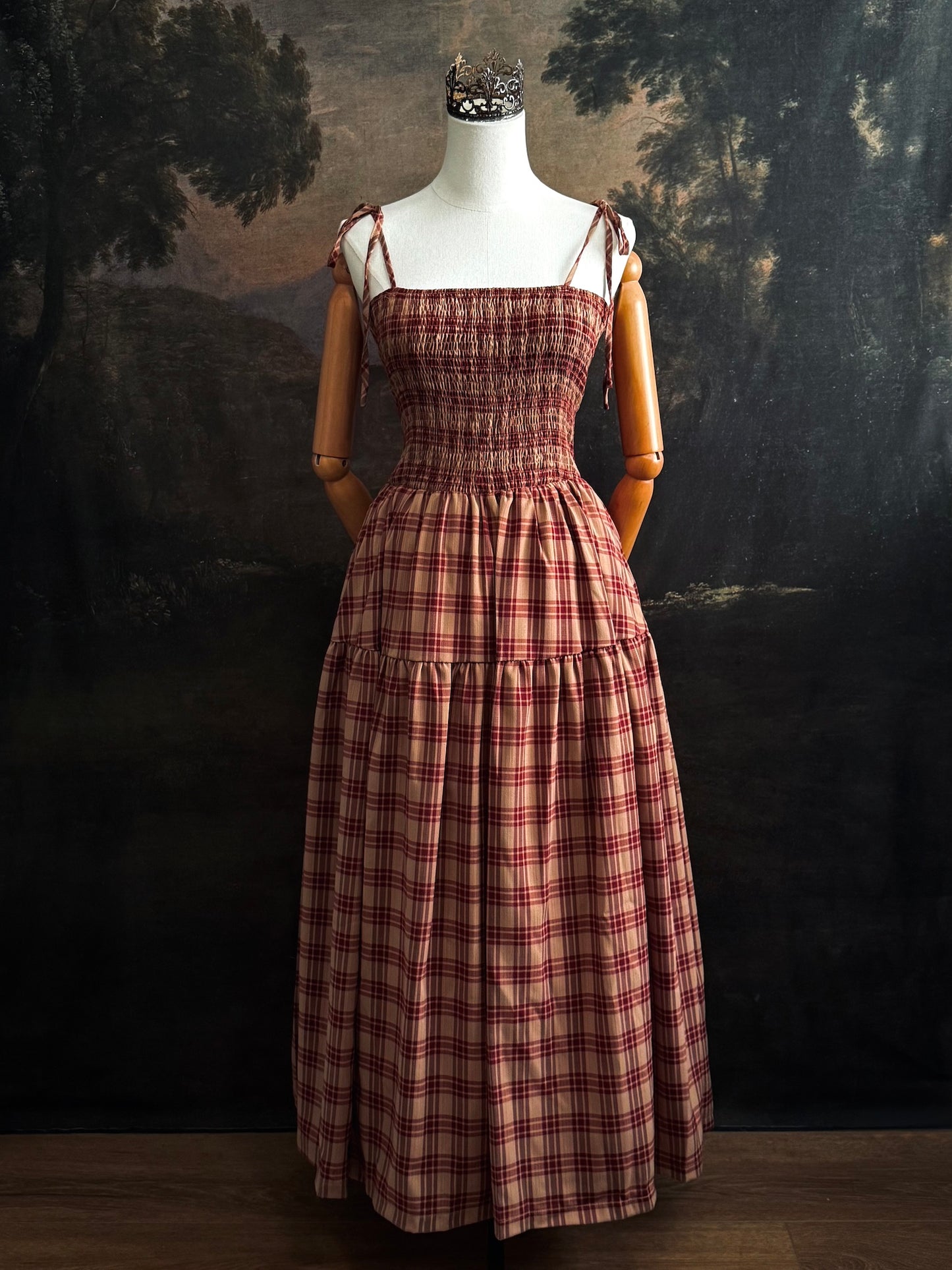A historically-inspired burgundy and tan highland tartan plaid maxi dress with tiered skirt, shirring, and self-tie straps. Pictured on a mannequin in front of a dark forested art history backdrop.
