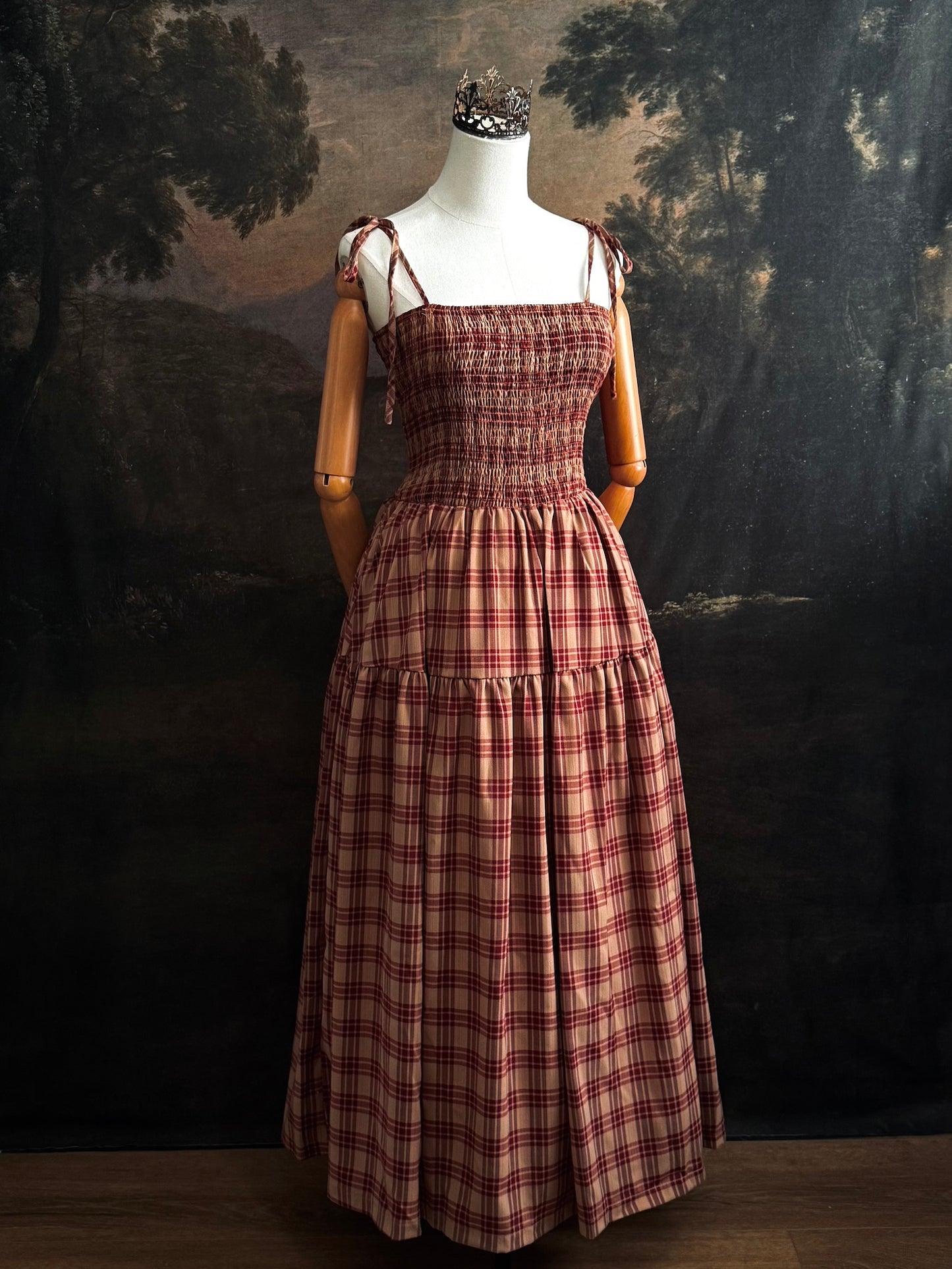 A historically-inspired burgundy and tan highland tartan plaid maxi dress with tiered skirt, shirring, and self-tie straps. Pictured on a mannequin in front of a dark forested art history backdrop.