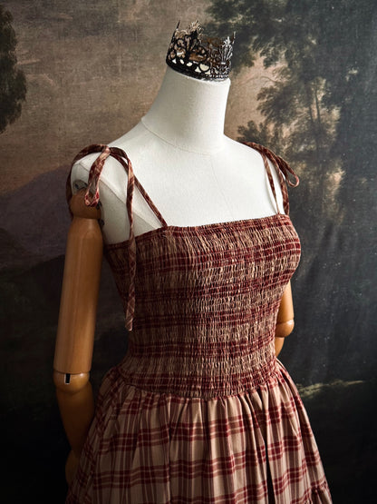 A historically-inspired burgundy and tan highland tartan plaid maxi dress with tiered skirt, shirring, and self-tie straps. Pictured on a mannequin in front of a dark forested art history backdrop.