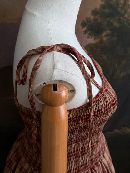 A historically-inspired burgundy and tan highland tartan plaid maxi dress with tiered skirt, shirring, and self-tie straps. Pictured on a mannequin in front of a dark forested art history backdrop.