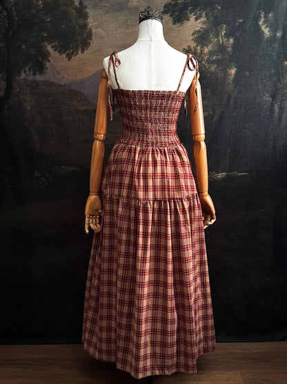 A historically-inspired burgundy and tan highland tartan plaid maxi dress with tiered skirt, shirring, and self-tie straps. Pictured on a mannequin in front of a dark forested art history backdrop.
