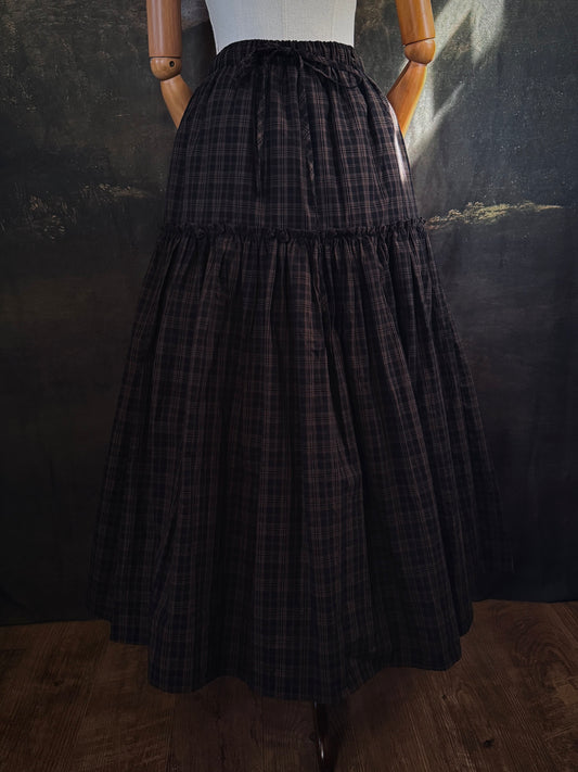 A long tiered cotton skirt in a dark brown and black plaid pattern, inspired by historical and vintage fashion, displayed on a mannequin against a dark historical landscape painting backdrop.