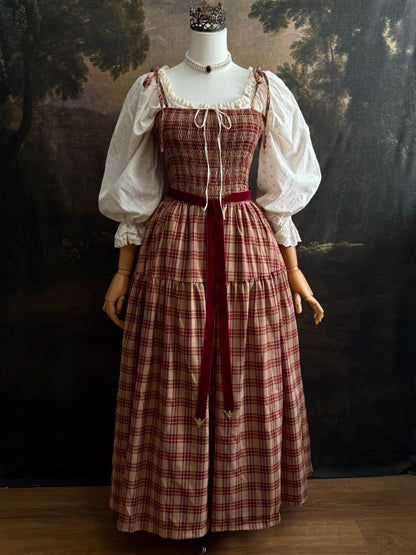 A medieval-style outfit displayed on a mannequin, featuring a smocked burgundy and beige plaid tartan maxi dress, an ivory cotton eyelet puff-sleeve chemise dress, and a dark red velvet sash.