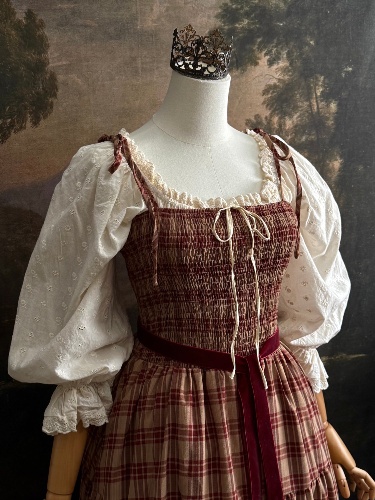 A medieval-style outfit displayed on a mannequin, featuring a smocked burgundy and beige plaid tartan maxi dress, an ivory cotton eyelet puff-sleeve chemise dress, and a dark red velvet sash.