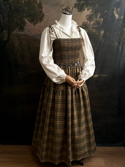 A mannequin dressed in a historical-style outfit featuring an ivory long-sleeve blouse layered under a smocked green tartan plaid dress, styled with a silver pendant necklace against a dark painted backdrop.