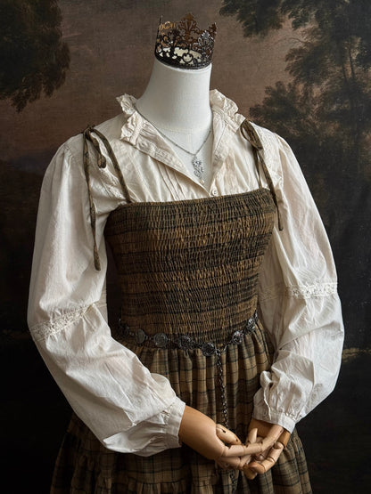 A mannequin dressed in a historical-style outfit featuring an ivory long-sleeve blouse layered under a smocked green tartan plaid dress, styled with a silver pendant necklace against a dark painted backdrop.