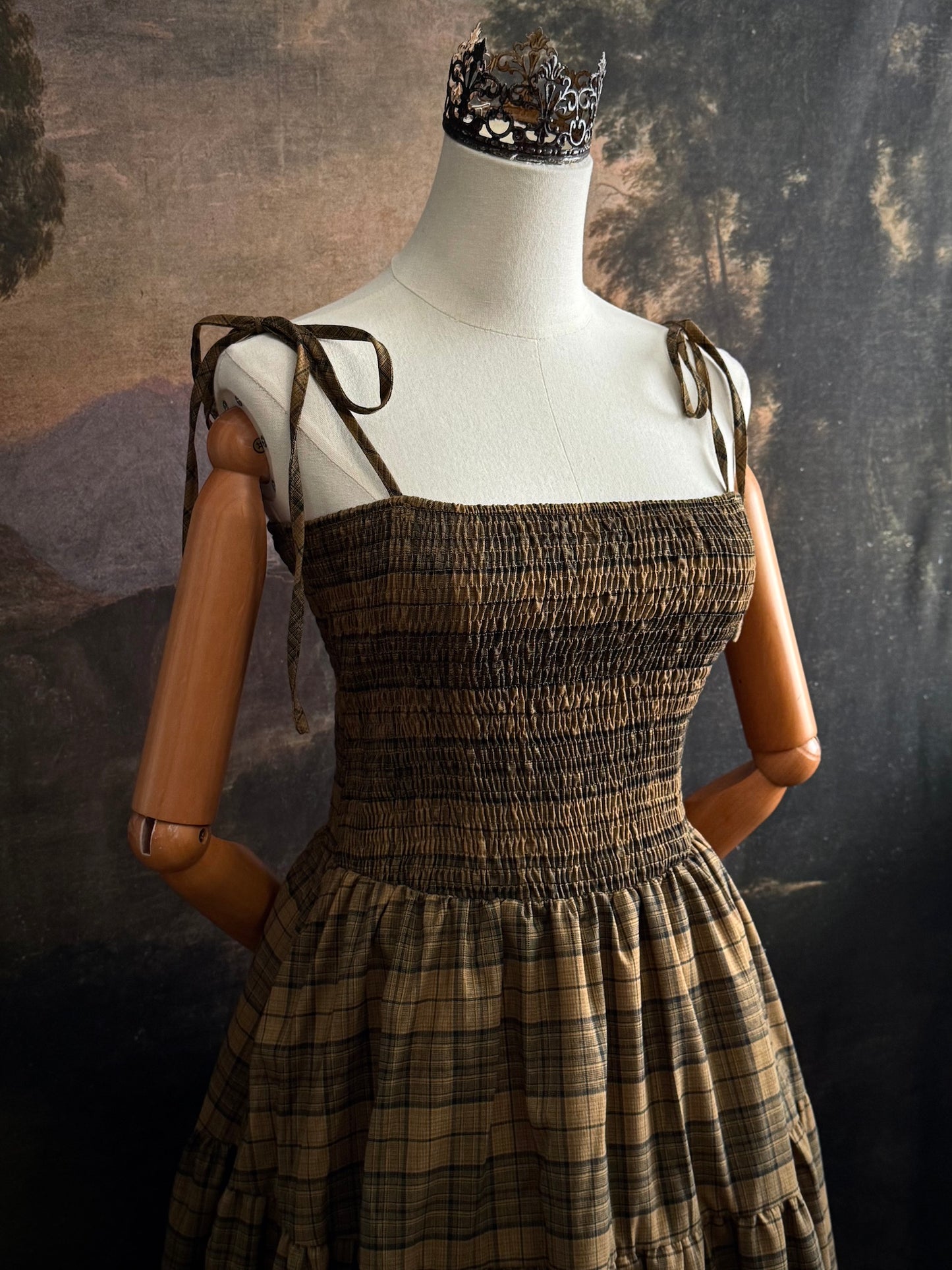 A historically-inspired celtic olive / moss green highland tartan plaid maxi dress with tiered skirt, shirring, and self-tie straps. Pictured on a mannequin in front of a dark forested art history backdrop.