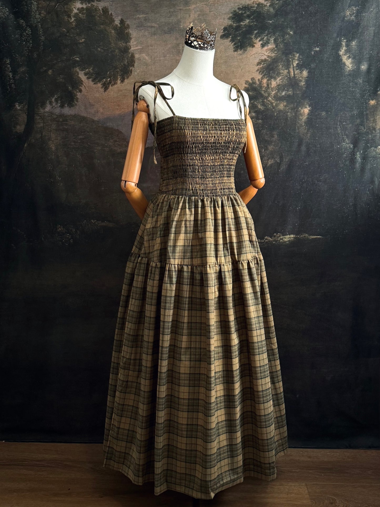 A historically-inspired celtic olive / moss green highland tartan plaid maxi dress with tiered skirt, shirring, and self-tie straps. Pictured on a mannequin in front of a dark forested art history backdrop.