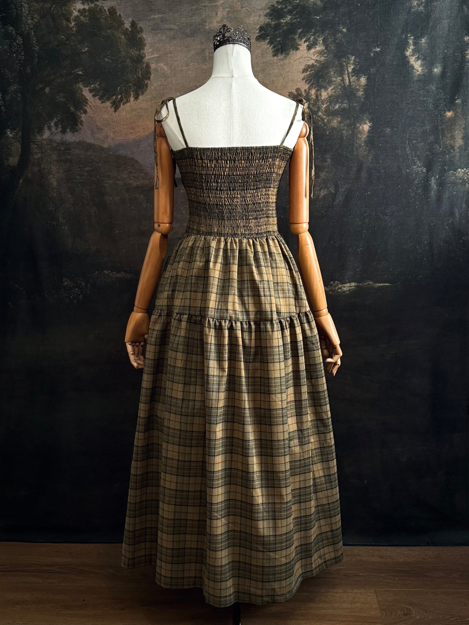 A historically-inspired celtic olive / moss green highland tartan plaid maxi dress with tiered skirt, shirring, and self-tie straps. Pictured on a mannequin in front of a dark forested art history backdrop.