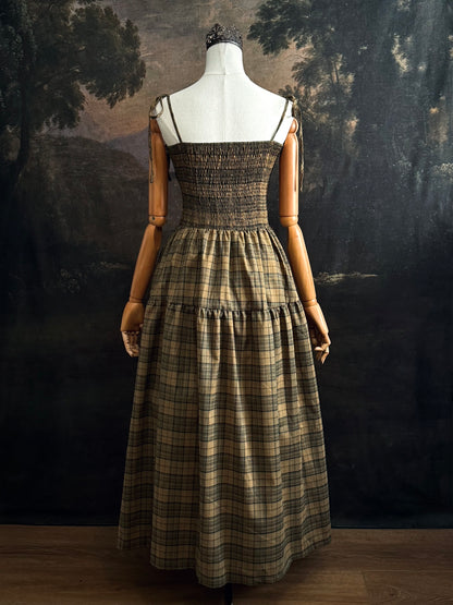 A historically-inspired celtic olive / moss green highland tartan plaid maxi dress with tiered skirt, shirring, and self-tie straps. Pictured on a mannequin in front of a dark forested art history backdrop.