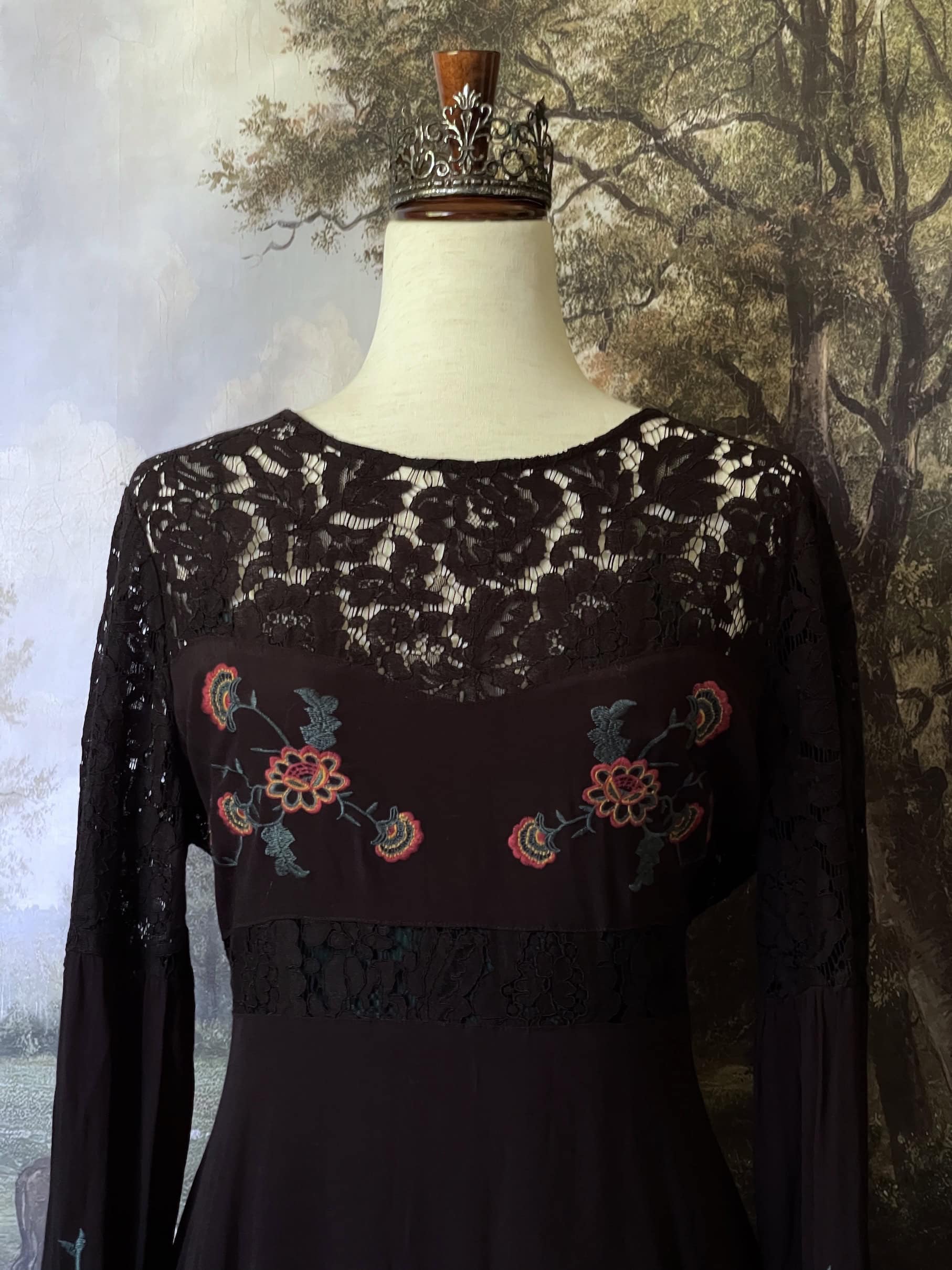 A black Historically-inspired western folk embroidered floral lace inset dress with bishop sleeves is pictured on a mannequin in front of an art history backdrop.