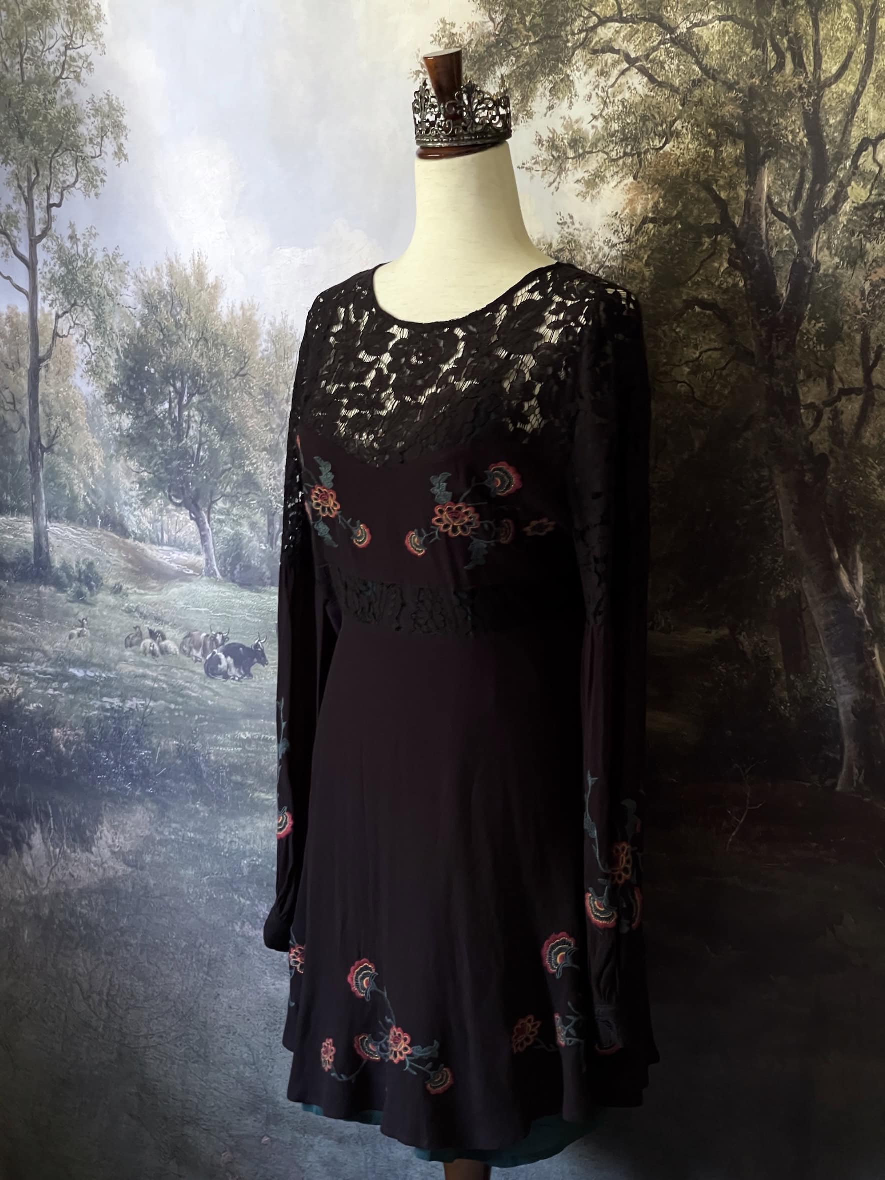 A black Historically-inspired western folk embroidered floral lace inset dress with bishop sleeves is pictured on a mannequin in front of an art history backdrop.