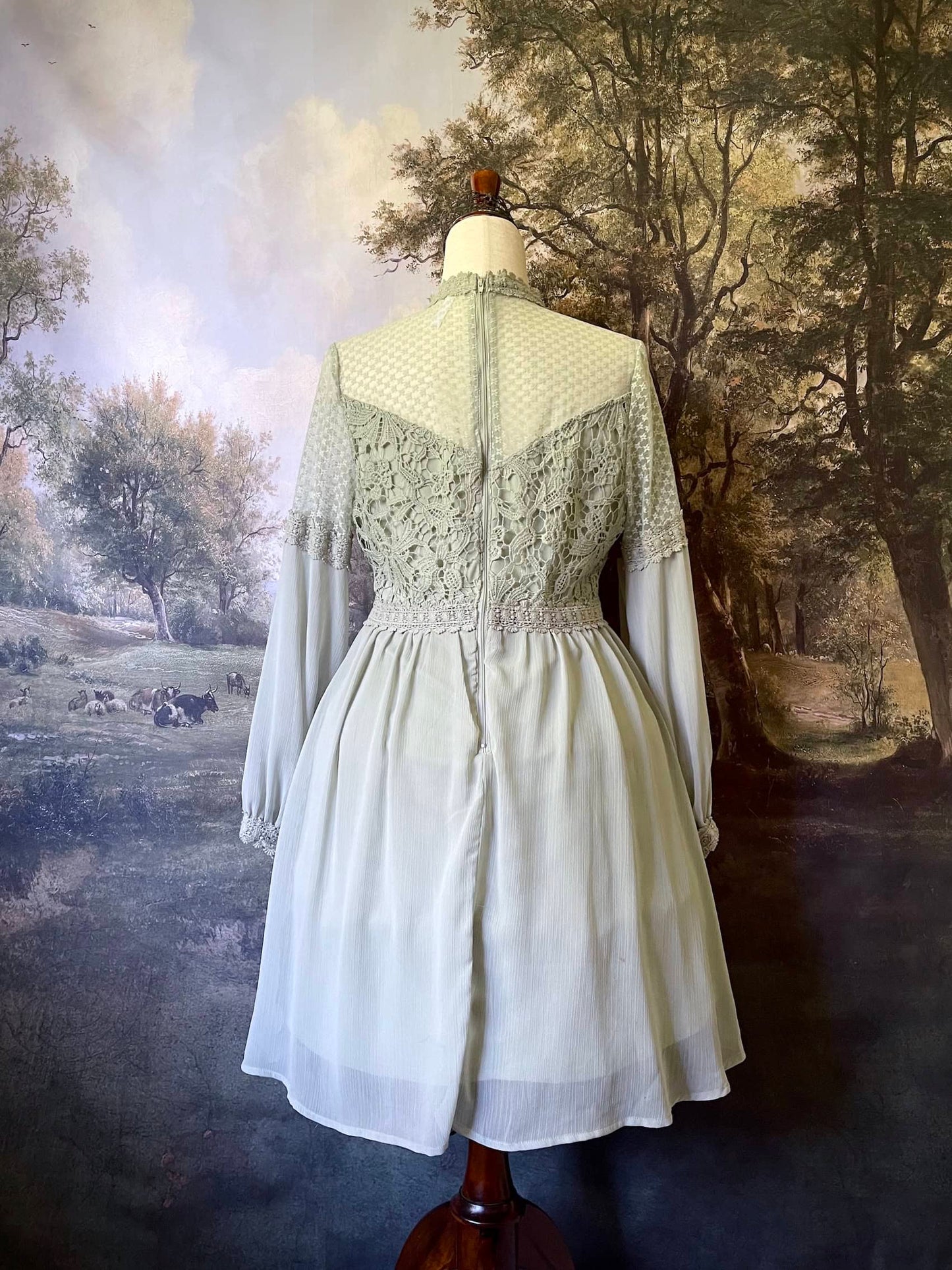 A historically inspired sage green edwardian fairytale dress with bishop sleeves and lace trim bodice is pictured on a mannequin in front of an art history backdrop.