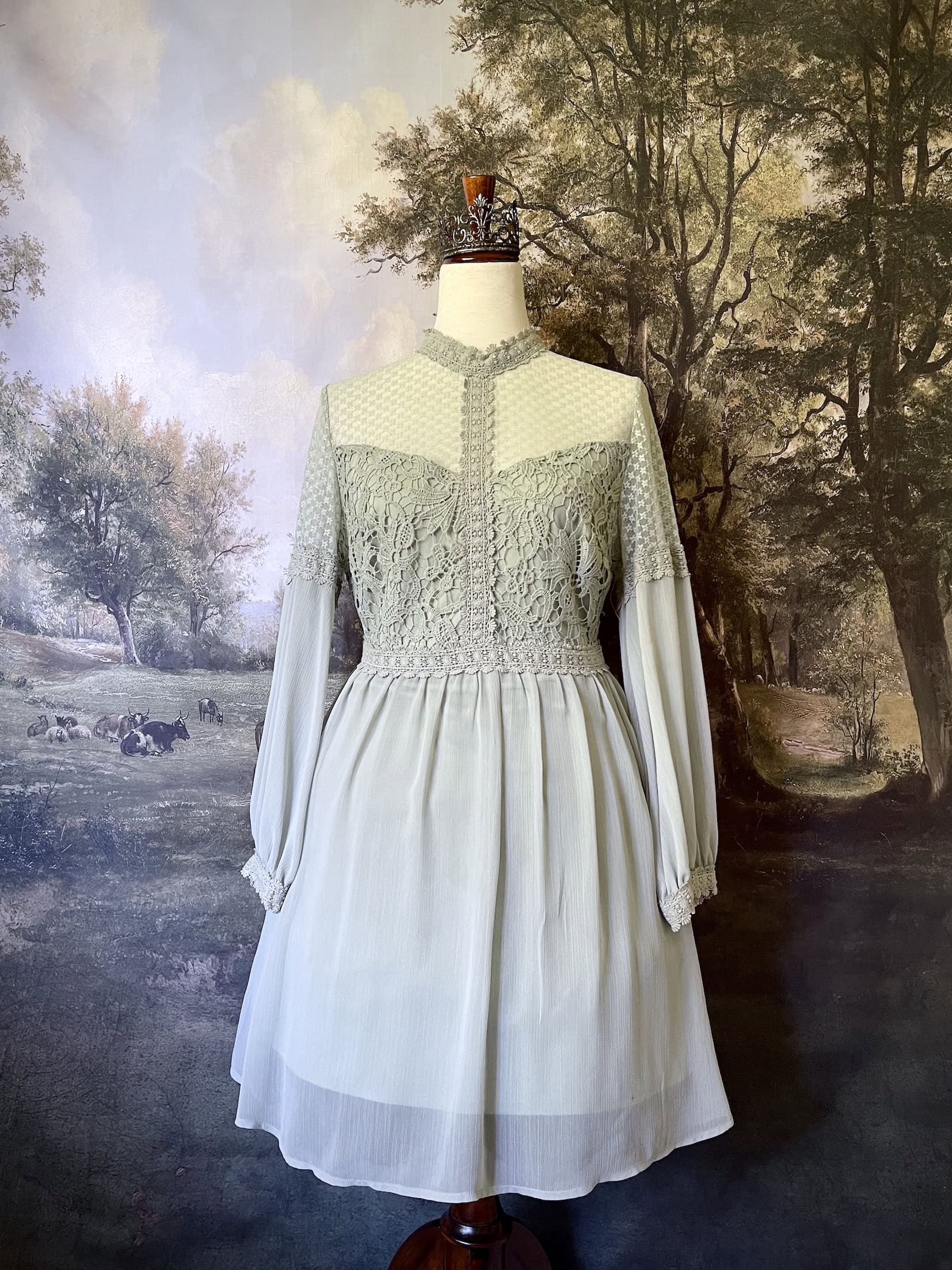 A historically inspired sage green edwardian fairytale dress with bishop sleeves and lace trim bodice is pictured on a mannequin in front of an art history backdrop.