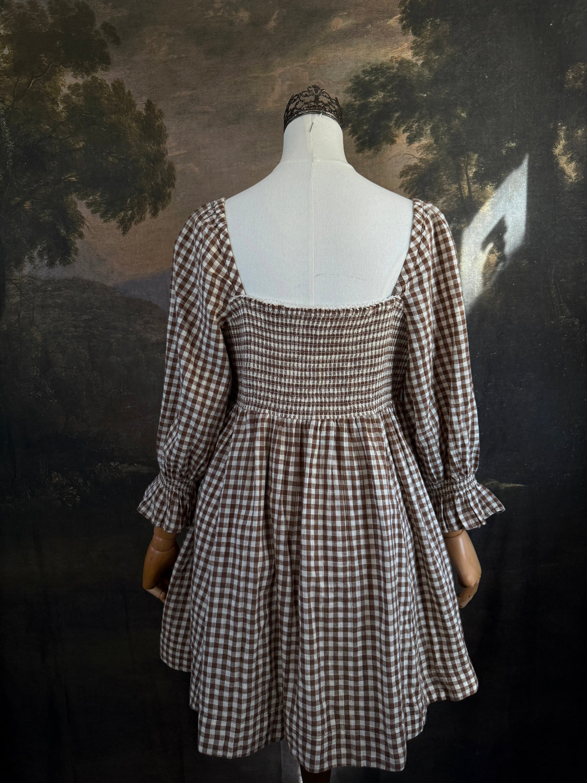 A vintage-inspired autumnal brown and white gingham check print long-sleeve mini dress with shirring and button-front, displayed on a mannequin against a dark classical landscape painting backdrop.