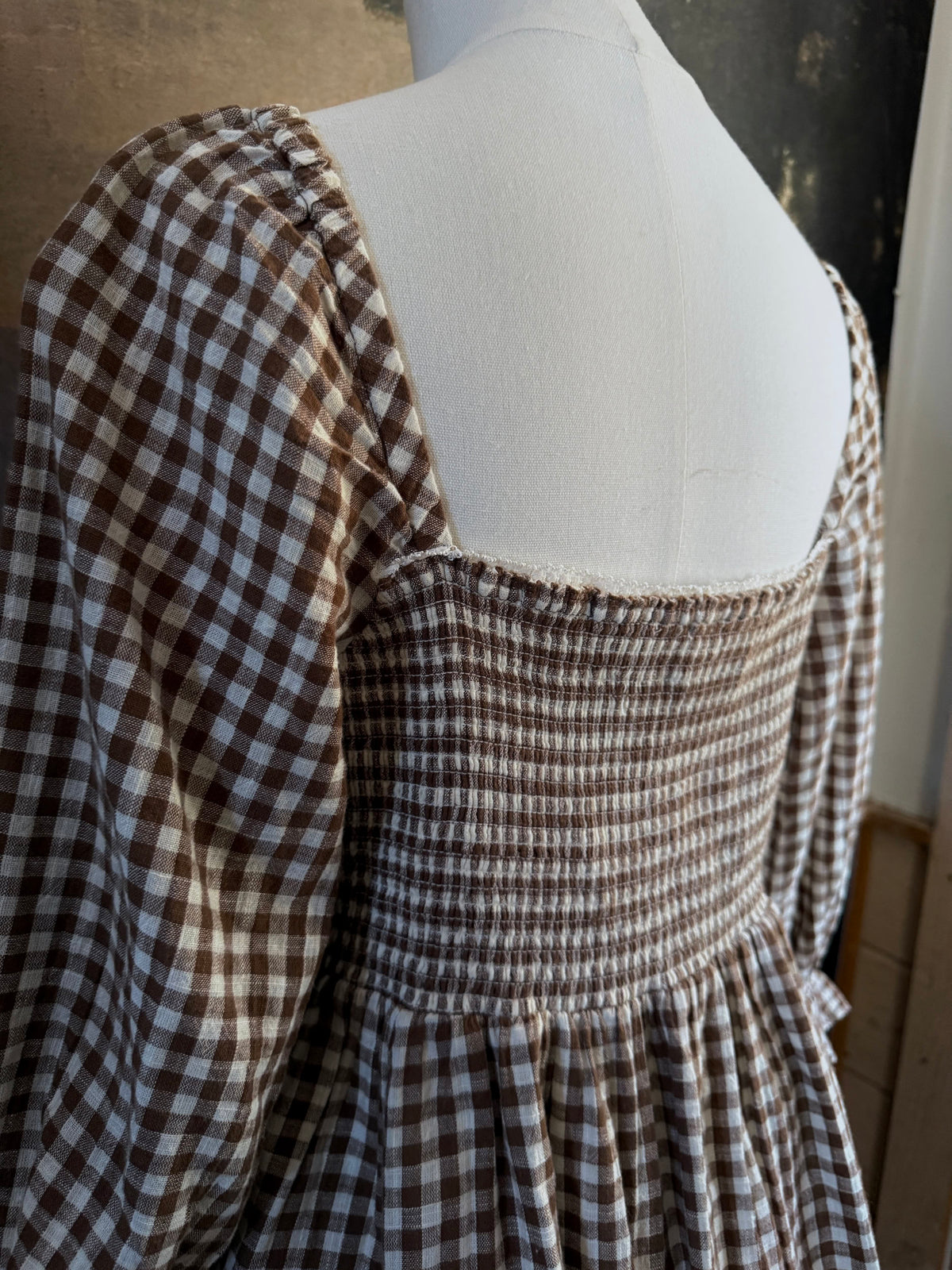 A vintage-inspired autumnal brown and white gingham check print long-sleeve mini dress with shirring and button-front, displayed on a mannequin against a dark classical landscape painting backdrop.