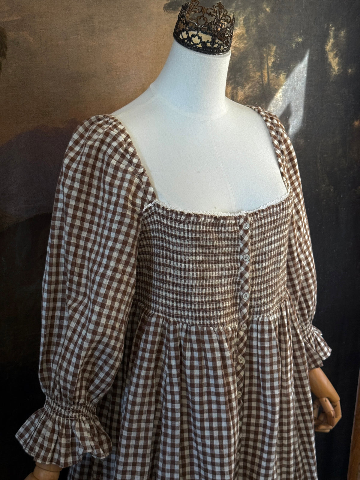 A vintage-inspired autumnal brown and white gingham check print long-sleeve mini dress with shirring and button-front, displayed on a mannequin against a dark classical landscape painting backdrop.