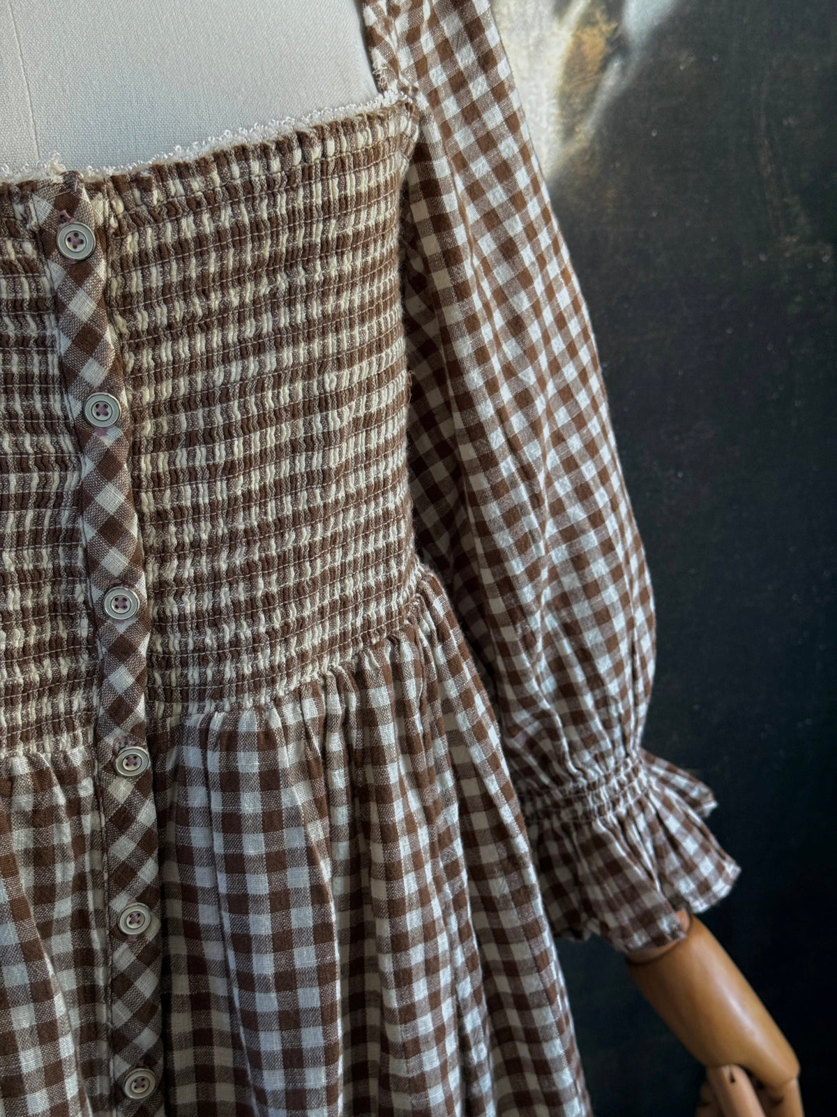 A vintage-inspired autumnal brown and white gingham check print long-sleeve mini dress with shirring and button-front, displayed on a mannequin against a dark classical landscape painting backdrop.
