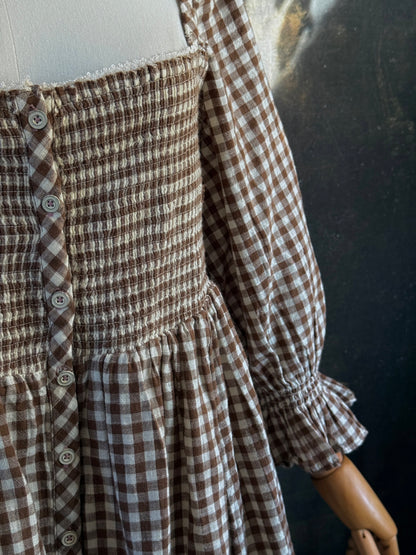 A vintage-inspired autumnal brown and white gingham check print long-sleeve mini dress with shirring and button-front, displayed on a mannequin against a dark classical landscape painting backdrop.