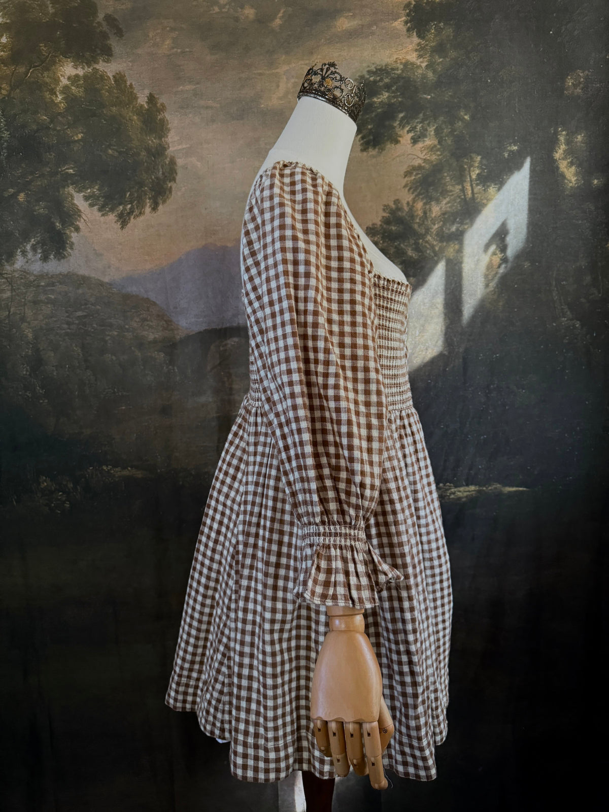 A vintage-inspired autumnal brown and white gingham check print long-sleeve mini dress with shirring and button-front, displayed on a mannequin against a dark classical landscape painting backdrop.