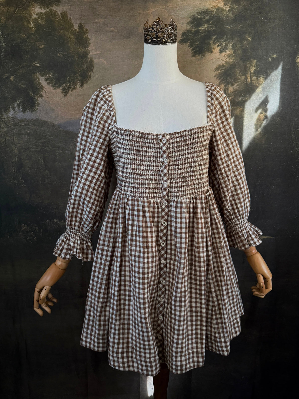 A vintage-inspired autumnal brown and white gingham check print long-sleeve mini dress with shirring and button-front, displayed on a mannequin against a dark classical landscape painting backdrop.