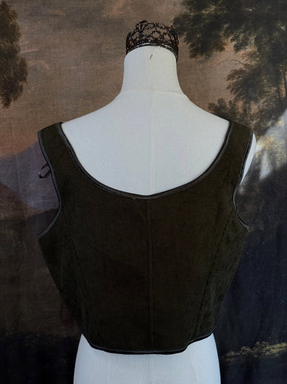 A historically inspired dark olive moss green corduroy corset top with brown lace-up front ties and shoulder ties, displayed on a mannequin against a scenic landscape painting backdrop.