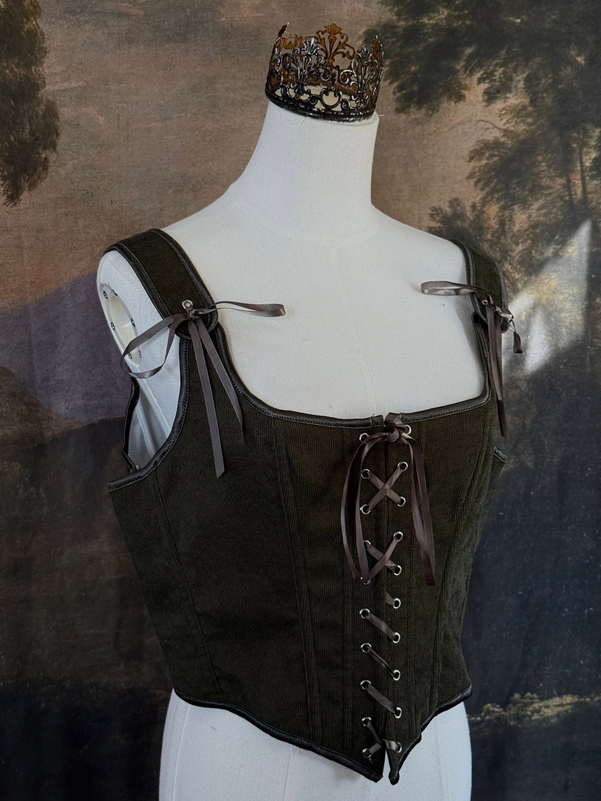 A historically inspired dark olive moss green corduroy corset top with brown lace-up front ties and shoulder ties, displayed on a mannequin against a scenic landscape painting backdrop.