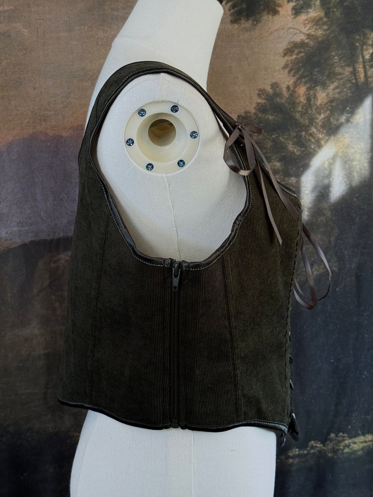 A historically inspired dark olive moss green corduroy corset top with brown lace-up front ties and shoulder ties, displayed on a mannequin against a scenic landscape painting backdrop.