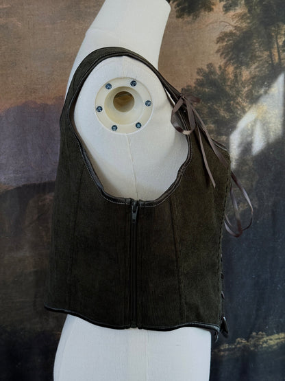 A historically inspired dark olive moss green corduroy corset top with brown lace-up front ties and shoulder ties, displayed on a mannequin against a scenic landscape painting backdrop.