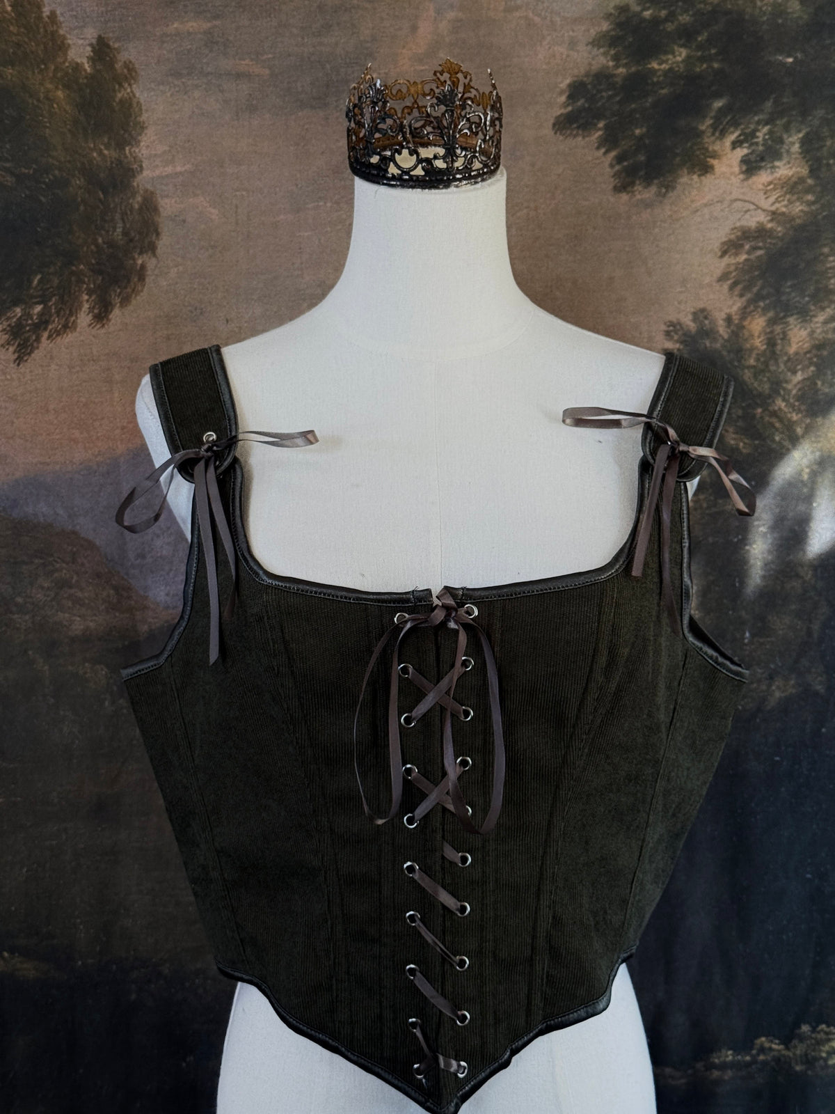 A historically inspired dark olive moss green corduroy corset top with brown lace-up front ties and shoulder ties, displayed on a mannequin against a scenic landscape painting backdrop.