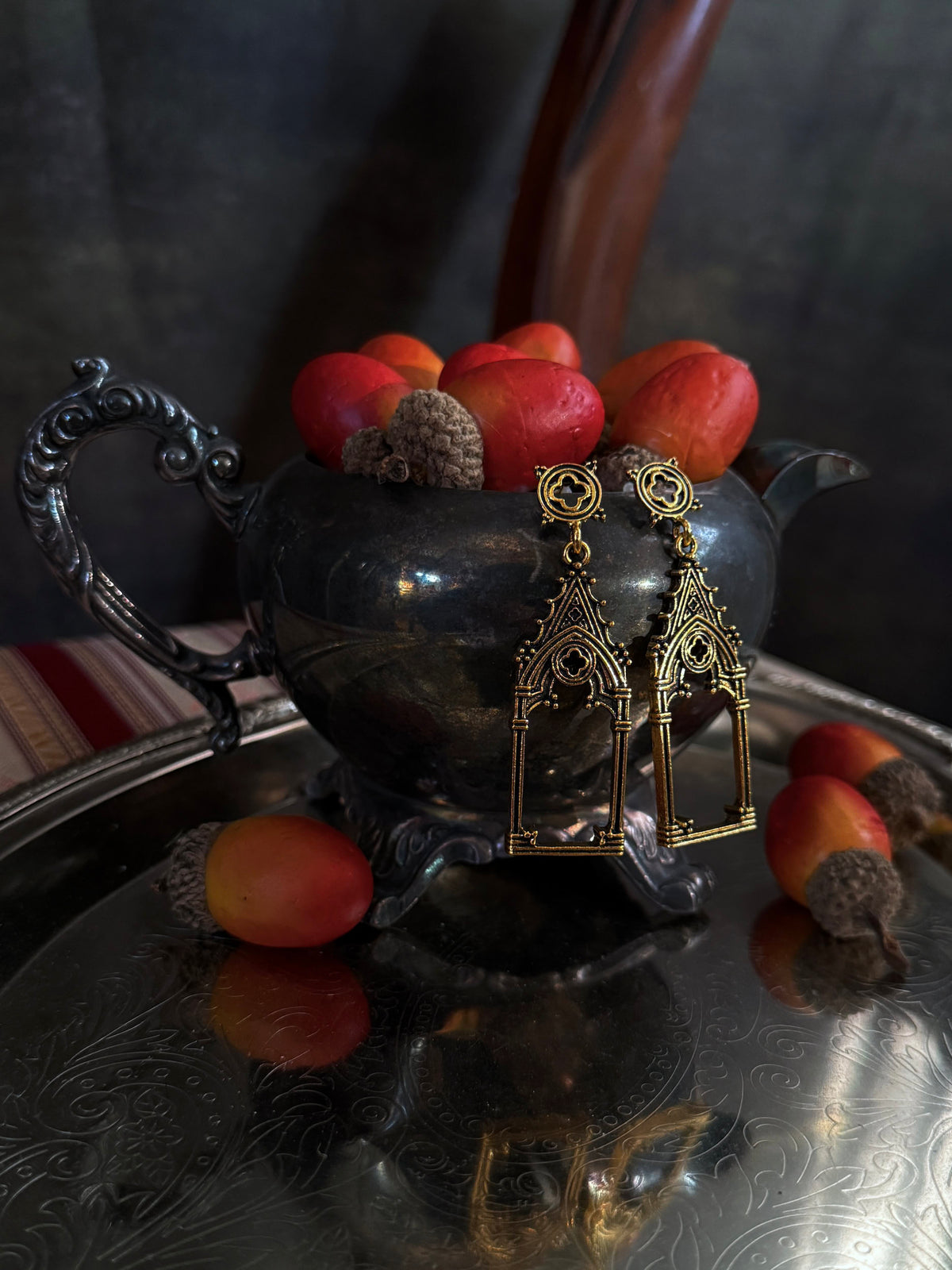 A pair of gold-toned gothic cathedral arch drop earrings with quatrefoil filigree studs, displayed on a dark metallic teapot filled with acorns on an etched metal tray.