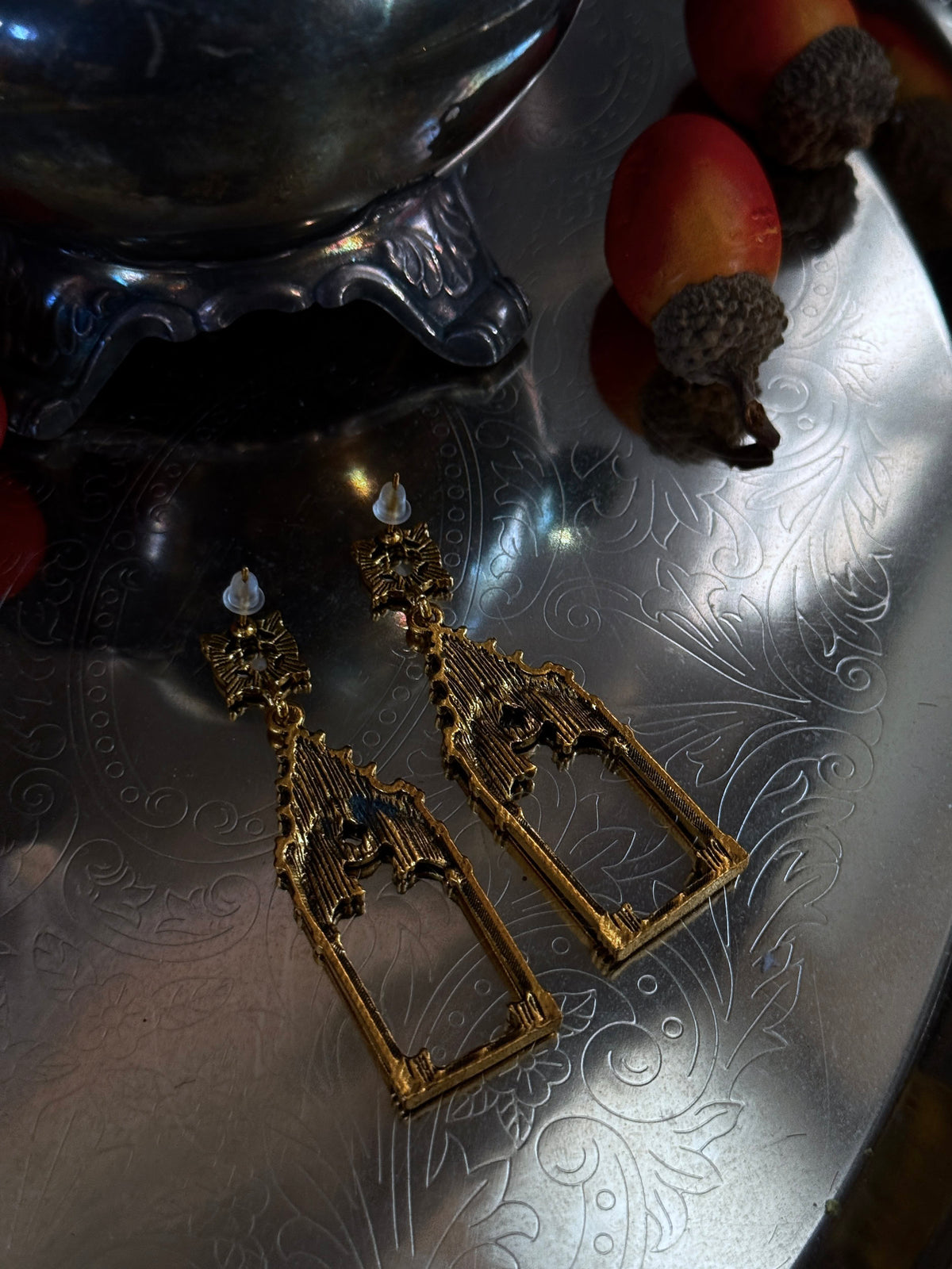 A pair of gold-toned gothic cathedral arch drop earrings with quatrefoil filigree studs, displayed on an etched metal tray.