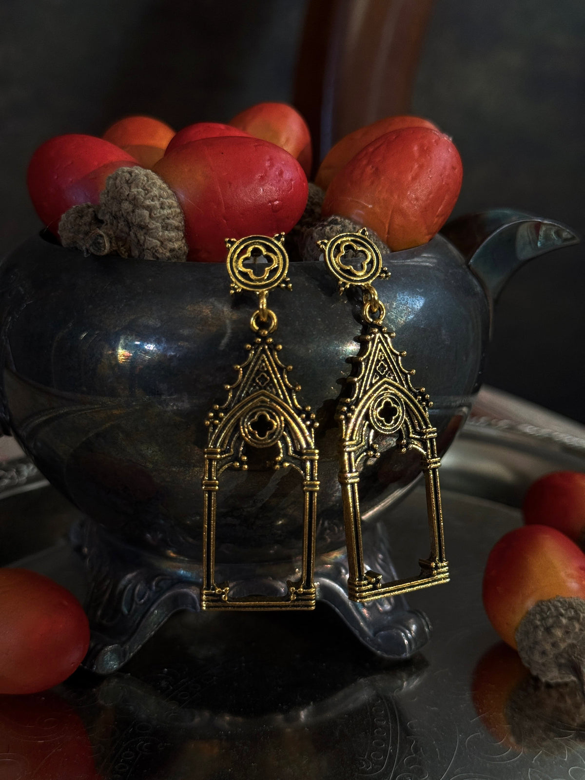A pair of gold-toned gothic cathedral arch drop earrings with quatrefoil filigree studs, displayed on a dark metallic teapot filled with acorns on an etched metal tray.