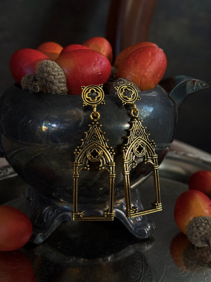A pair of gold-toned gothic cathedral arch drop earrings with quatrefoil filigree studs, displayed on a dark metallic teapot filled with acorns on an etched metal tray.