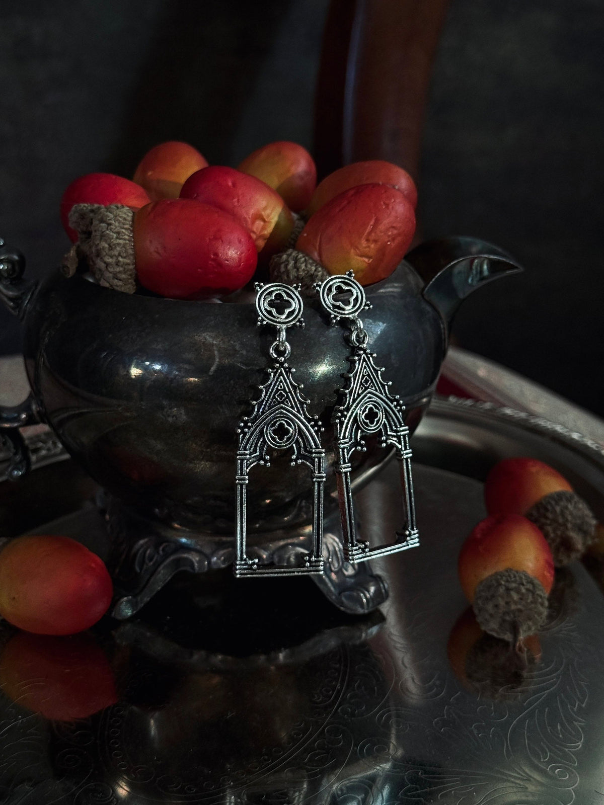A pair of silver medievak inspired gothic arch drop dangke earrings, hanging from an antique pewter teapot filled with acorns, set on a reflective silver tray.