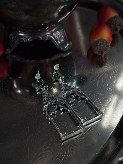 A pair of medieval inspired silver-toned gothic cathedral arch earrings resting on a polished silver metal tray, with decorative red acorns in the background.