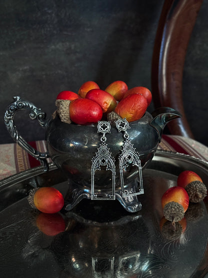 A pair of silver medievak inspired gothic arch drop dangke earrings, hanging from an antique pewter teapot filled with acorns, set on a reflective silver tray.