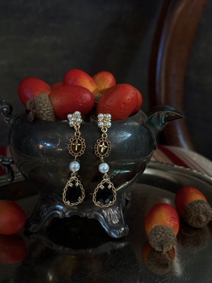 A pair of renaissance inspired ornate gold and black drop earrings with white pearl-like beads, hanging from a dark antique  pot filled with red acorns on a reflective silver tray.