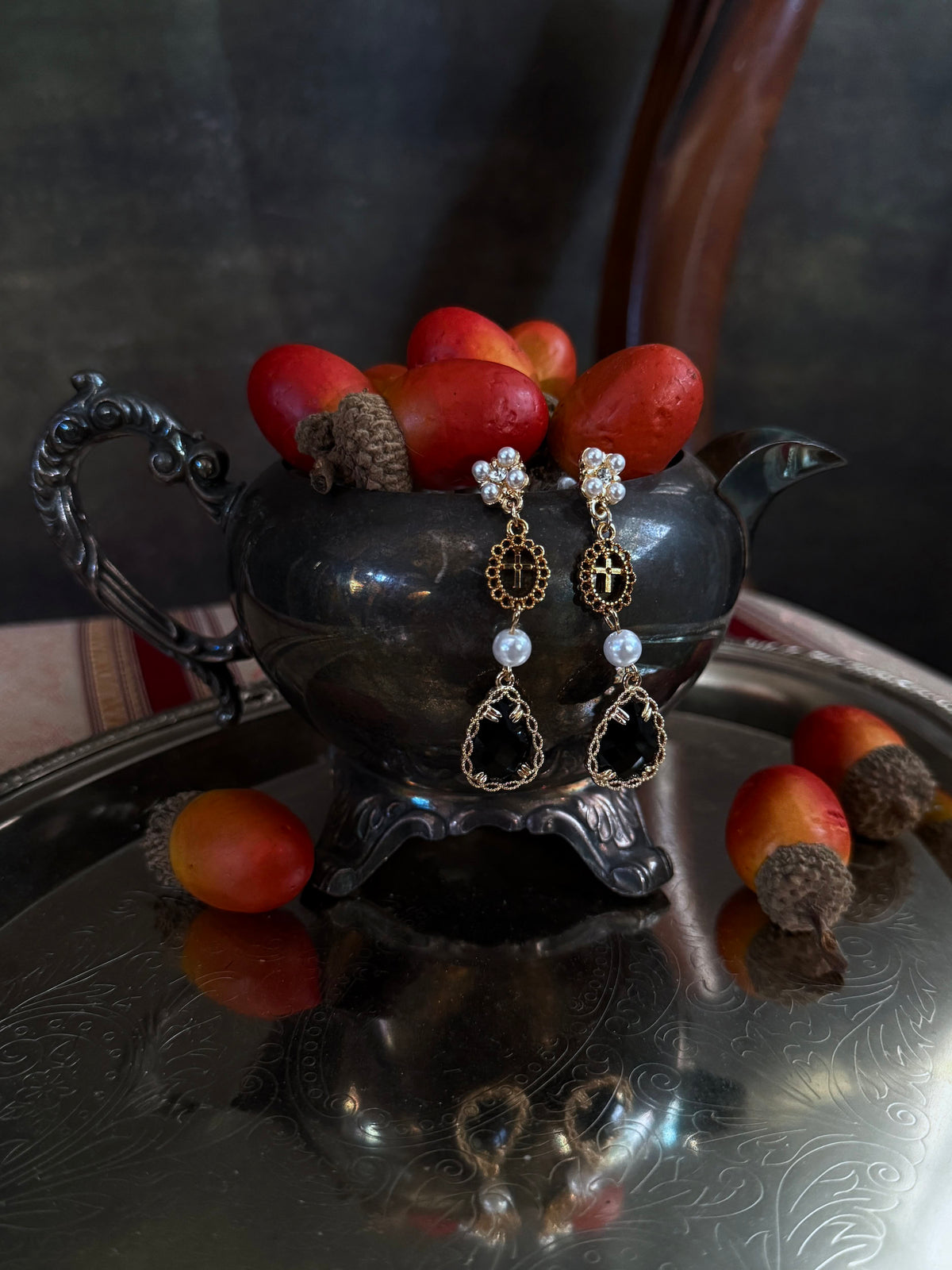 A pair of renaissance inspired ornate gold and black drop earrings with white pearl-like beads, hanging from a dark antique  pot filled with red acorns on a reflective silver tray.