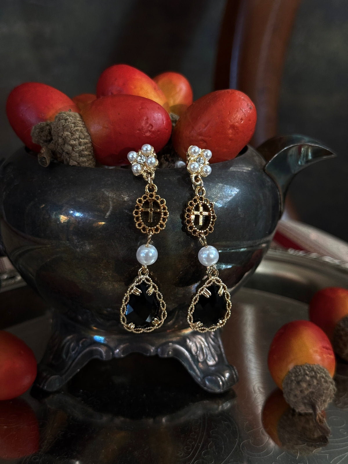 A pair of renaissance inspired ornate gold and black drop earrings with white pearl-like beads, hanging from a dark antique  pot filled with red acorns on a reflective silver tray.