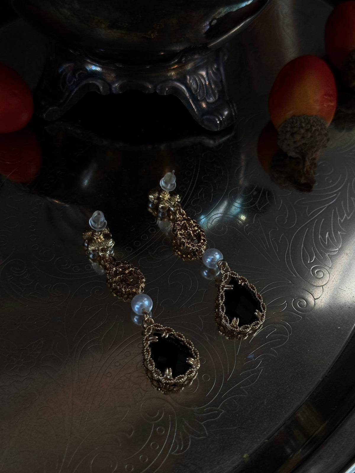 A pair of renaissance inspired ornate gold and black drop earrings with white pearl-like beads, hanging from a dark antique  pot filled with red acorns on a reflective silver tray.