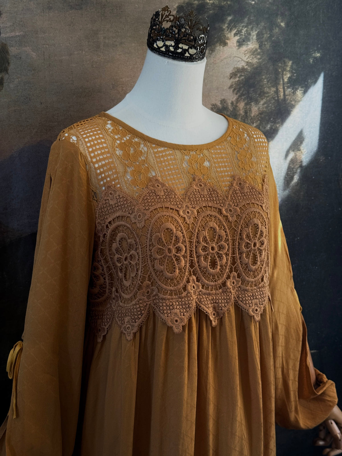 A medieval renaissance inspired tunic dress with cathedral arch lace, billowy slashed bishop sleeves, all in a golden yellow fabric. Pictured on a mannequin in front of a historical painting backdrop. Close up.